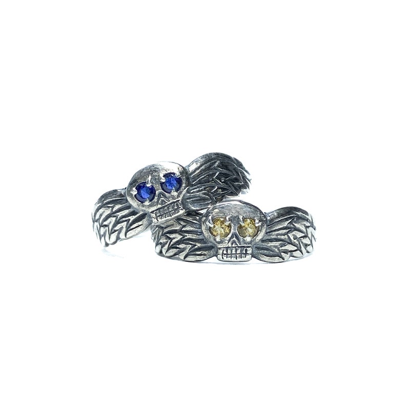 Puede incluir: Dos anillos de plata con forma de calavera y alas. Una calavera tiene ojos azules y la otra tiene ojos amarillos.