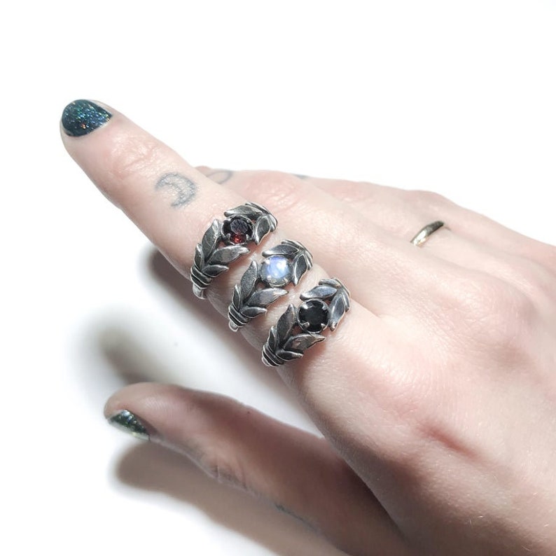 May include: Three silver rings with leaf designs and different coloured gemstones. The rings are stacked on a finger.
