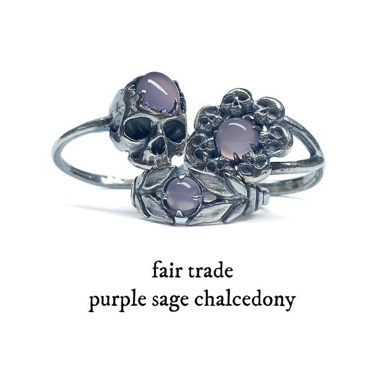 May include: Three silver rings featuring purple sage chalcedony stones. The rings depict a skull, a flower with skulls, and a leafy design. The rings are labelled "fair trade".
