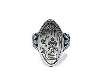 Puede incluir: Un anillo de plata con un diseño ovalado que presenta una imagen grabada de un jarrón con un árbol detrás.