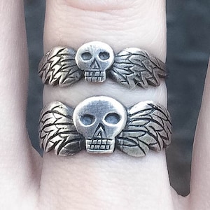 May include: Two silver rings with a skull design and wings. The wings are detailed and the skulls have hollow eye sockets.
