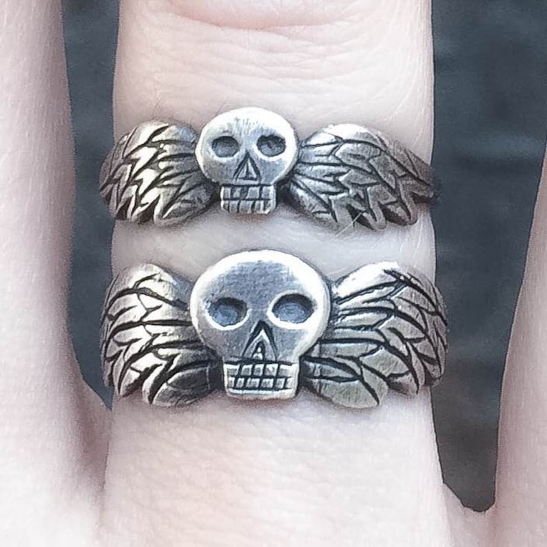 May include: Two silver rings with a skull design and wings. The wings are detailed and the skulls have hollow eye sockets.