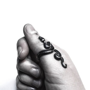 May include: A silver snake ring with a detailed design. The ring is worn on the thumb of a hand.