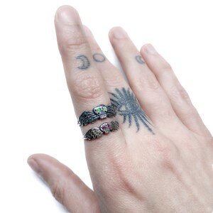 Puede incluir: Dos anillos de plata con forma de calavera y alas. El anillo superior tiene una piedra verde para los ojos y el anillo inferior tiene piedras rosas para los ojos.
