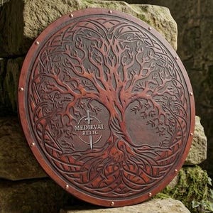 Może przedstawiać: Okrągła tarcza z brązowej skóry z wytłoczonym wzorem drzewa o skomplikowanych korzeniach i gałęziach. Tekst MEDIEVAL RELIC jest wyśrodkowany nad ikoną miecza. Tarcza ma gładką krawędź zabezpieczoną małymi metalowymi nitami na obwodzie.
