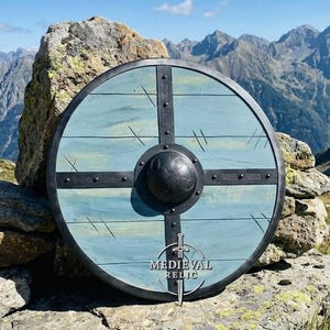 May include: A round wooden shield with a light blue finish and a black metal cross frame. The shield features a central black metal dome, dark metal rivets, and a black rim. The text "MEDIEVAL RELIC" is printed in white near the bottom center.
