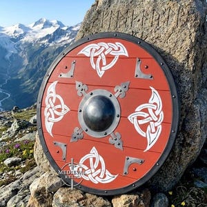 May include: A round wooden shield painted in a deep red hue, featuring white Celtic knotwork patterns, a central dark metal boss, and four decorative metal corner plates. The shield displays the text MEDIEVAL RELIC near the bottom edge.