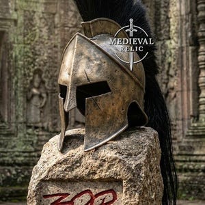 May include: A bronze-coloured Spartan-style helmet with a tall black crest, featuring a weathered finish and a T-shaped face opening. The helmet is displayed on a stone block with the red text 300 and a circular logo reading MEDIEVAL RELIC.