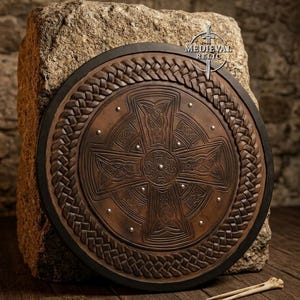 Könnte beinhalten: Ein runder Schild aus braunem Leder mit einem geprägten keltischen Kreuz in der Mitte, umgeben von einem geflochtenen Lederrand und verziert mit kleinen silberfarbenen Metallnieten. Der Text MEDIEVAL RELIC ist oben rechts zu sehen.