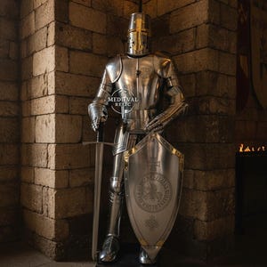 May include: A full suit of polished steel armor, including a helmet, breastplate, and leg guards, stands in a stone room. A sword rests beside the armor, and a shield with a crest is held in place. The armor is from Medieval Relic.