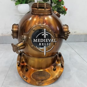 May include: A vintage brass diving helmet with a round, porthole-like window displaying the text "MEDIEVAL RELIC" and a sword emblem. The helmet has a circular base and multiple fittings.