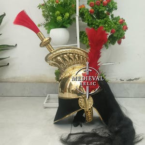 Op de afbeelding: Een goudkleurige helm met een zwarte rand en een rode pluim. De helm heeft ingewikkelde details en een lange zwarte paardenhaarpluim. De tekst "MEDIEVAL RELIC" is zichtbaar op de voorkant van de helm.