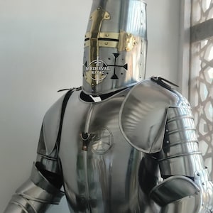 May include: A polished steel suit of armor, including a helmet with brass accents and a cross design. The armor features articulated plates for the arms and legs, and a chest plate with a cross emblem. The text "MEDIEVAL RELIC" is visible on the helmet.