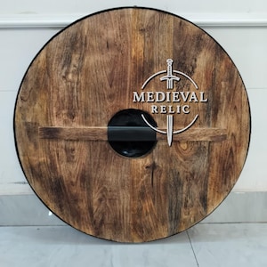 Puede incluir: Un escudo redondo de madera con acabado marr&oacute;n oscuro y un asa negra. El escudo presenta el texto "MEDIEVAL RELIC" en blanco, junto con un dise&ntilde;o de espada. Es un art&iacute;culo decorativo.