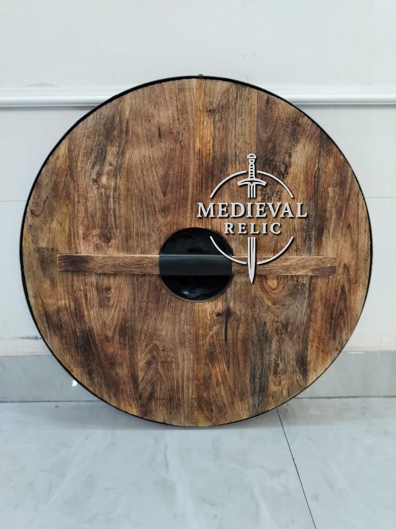 Puede incluir: Un escudo redondo de madera con acabado marr&oacute;n oscuro y un asa negra. El escudo presenta el texto "MEDIEVAL RELIC" en blanco, junto con un dise&ntilde;o de espada. Es un art&iacute;culo decorativo.