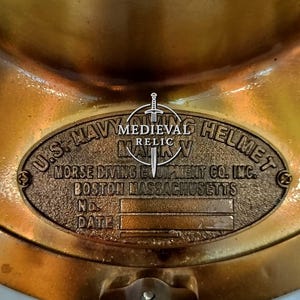 May include: A close-up of a vintage brass U.S. Navy diving helmet. The helmet features an oval plaque with embossed text: "U.S. NAVY DIVING HELMET MARK V" and "MORSE DIVING EQUIPMENT CO. INC. BOSTON MASSACHUSETTS". The helmet is a relic.