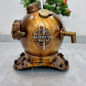 May include: A vintage brass diving helmet with the text "MEDIEVAL RELIC" on the front. The helmet has a round body, a circular top, and a base with multiple bolts. It features a spout and various other fittings, showcasing an antique aesthetic.