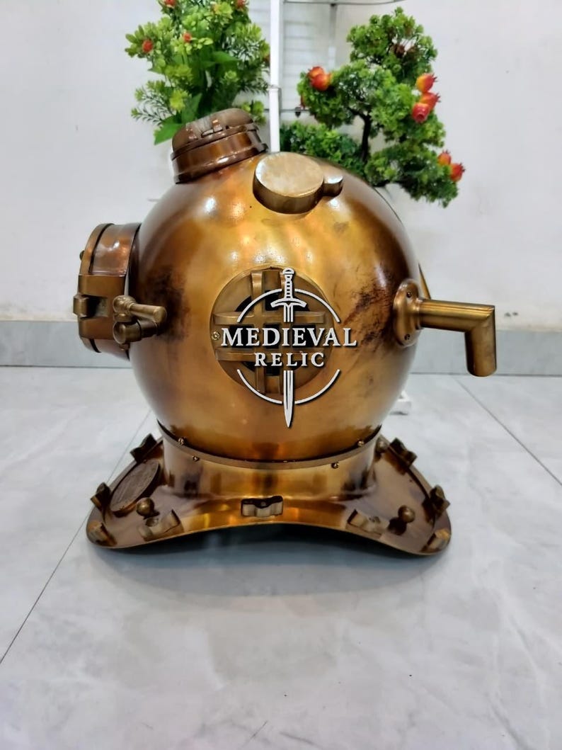 May include: A vintage brass diving helmet with the text "MEDIEVAL RELIC" on the front. The helmet has a round body, a circular top, and a base with multiple bolts. It features a spout and various other fittings, showcasing an antique aesthetic.