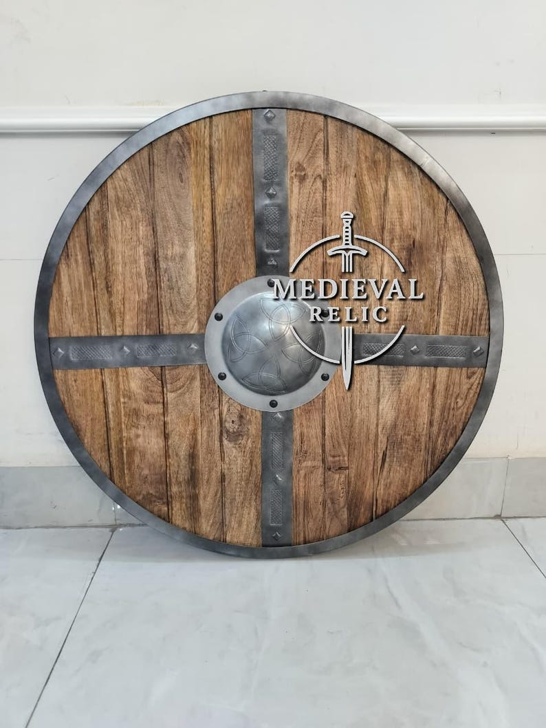 Puede incluir: Un escudo de madera redondo con borde y detalles met&aacute;licos. El escudo presenta un centro met&aacute;lico y la inscripci&oacute;n "MEDIEVAL RELIC". La madera tiene un veteado natural y es de color marr&oacute;n claro.