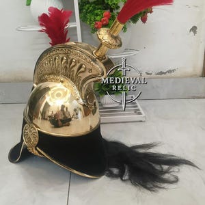 Op de afbeelding: Een goudkleurige metalen helm met een zwarte fluwelen basis en een lange zwarte pluim. De helm heeft sierlijke details en een rode pluim bovenop. De woorden "MEDIEVAL RELIC" staan op de zijkant.