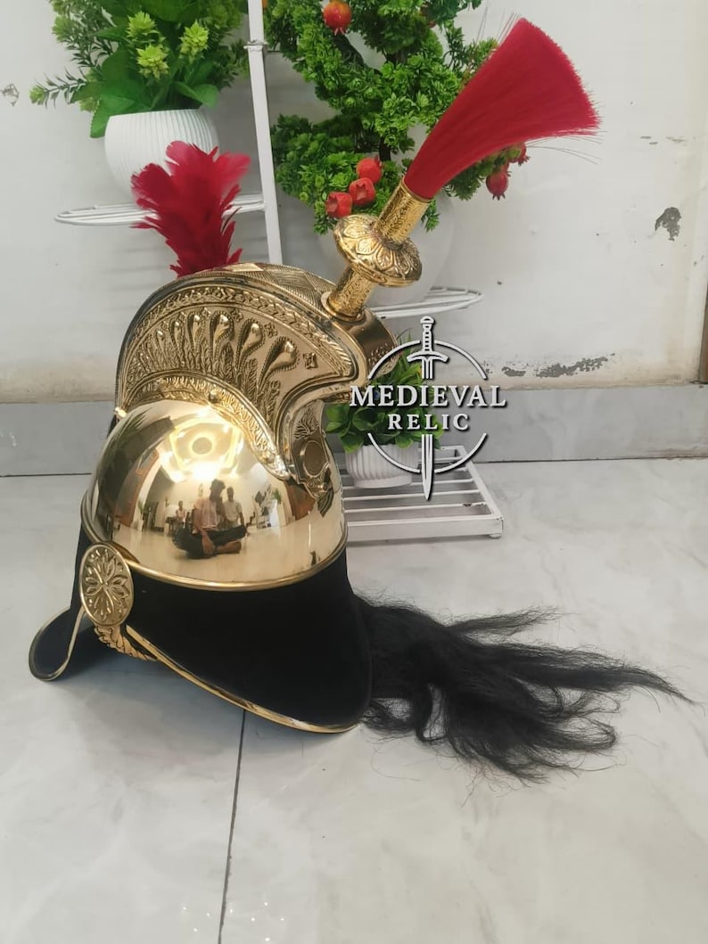 Op de afbeelding: Een goudkleurige metalen helm met een zwarte fluwelen basis en een lange zwarte pluim. De helm heeft sierlijke details en een rode pluim bovenop. De woorden "MEDIEVAL RELIC" staan op de zijkant.