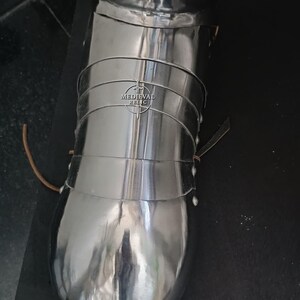May include: Polished metal medieval leg armor. The armor is made of multiple curved plates, riveted together. The image shows a close-up of the lower leg and foot protection, with the "Medieval Relic" logo visible.
