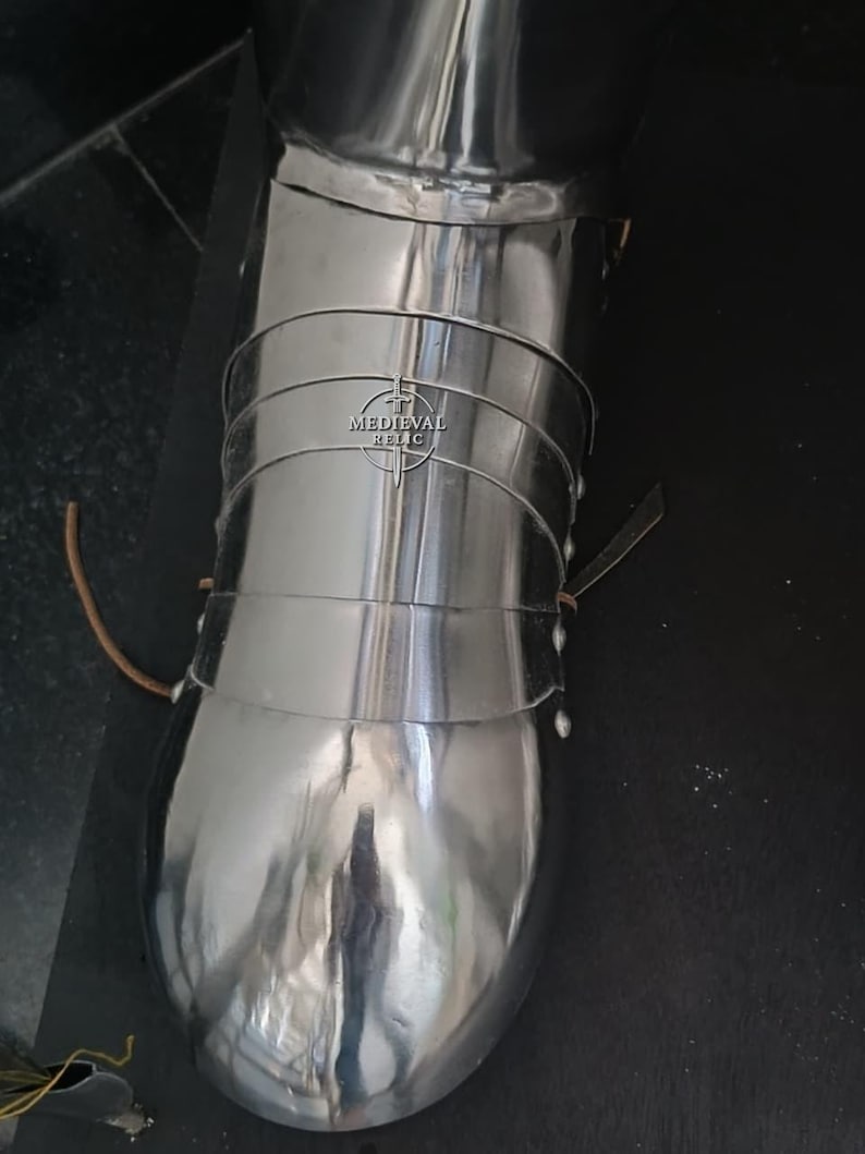 May include: Polished metal medieval leg armor. The armor is made of multiple curved plates, riveted together. The image shows a close-up of the lower leg and foot protection, with the "Medieval Relic" logo visible.