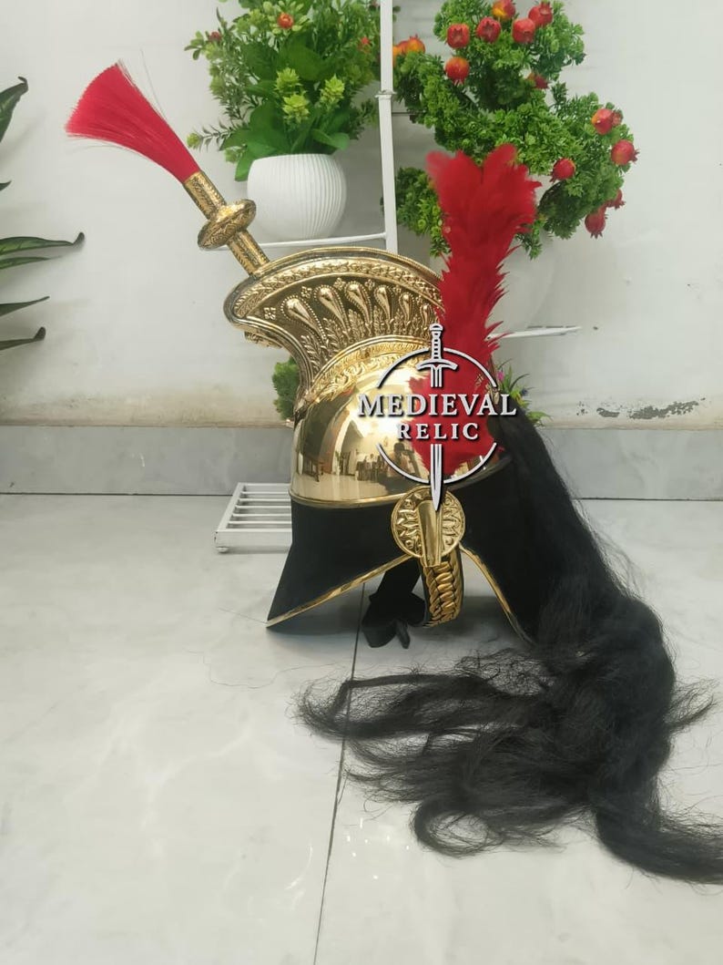 Op de afbeelding: Een goudkleurige helm met een zwarte rand en een rode pluim. De helm heeft ingewikkelde details en een lange zwarte paardenhaarpluim. De tekst "MEDIEVAL RELIC" is zichtbaar op de voorkant van de helm.