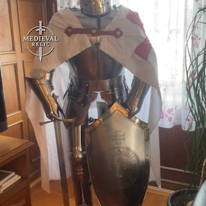 May include: A full suit of medieval armor, including a helmet, breastplate, and leg guards. The armor is made of polished metal and features a shield with a cross emblem. A white cape with a red cross is draped over the shoulders.