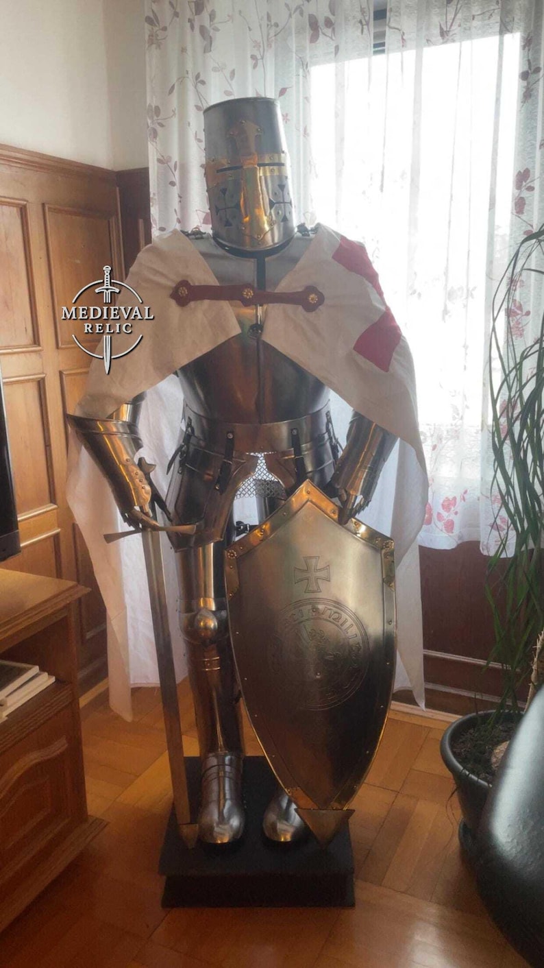 May include: A full suit of medieval armor, including a helmet, breastplate, and leg guards. The armor is made of polished metal and features a shield with a cross emblem. A white cape with a red cross is draped over the shoulders.