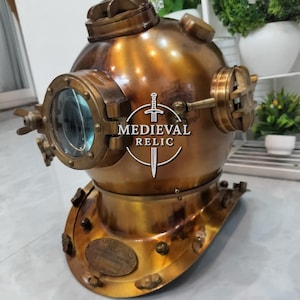 May include: A vintage brass diving helmet with a round, glass-covered viewport and multiple fittings. The helmet has a weathered, antique finish and a base with several bolts. The text "MEDIEVAL RELIC" is visible on the front.