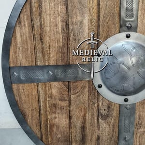 Puede incluir: Un escudo de madera redondo con borde y barras transversales de metal. El escudo tiene un jefe central de metal con un dise&ntilde;o de nudo celta. El texto "MEDIEVAL RELIC" est&aacute; impreso en el escudo.