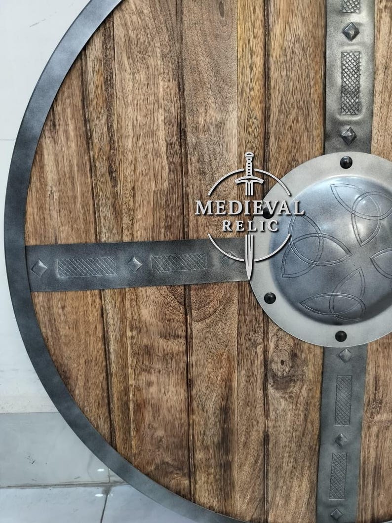 Puede incluir: Un escudo de madera redondo con borde y barras transversales de metal. El escudo tiene un jefe central de metal con un dise&ntilde;o de nudo celta. El texto "MEDIEVAL RELIC" est&aacute; impreso en el escudo.