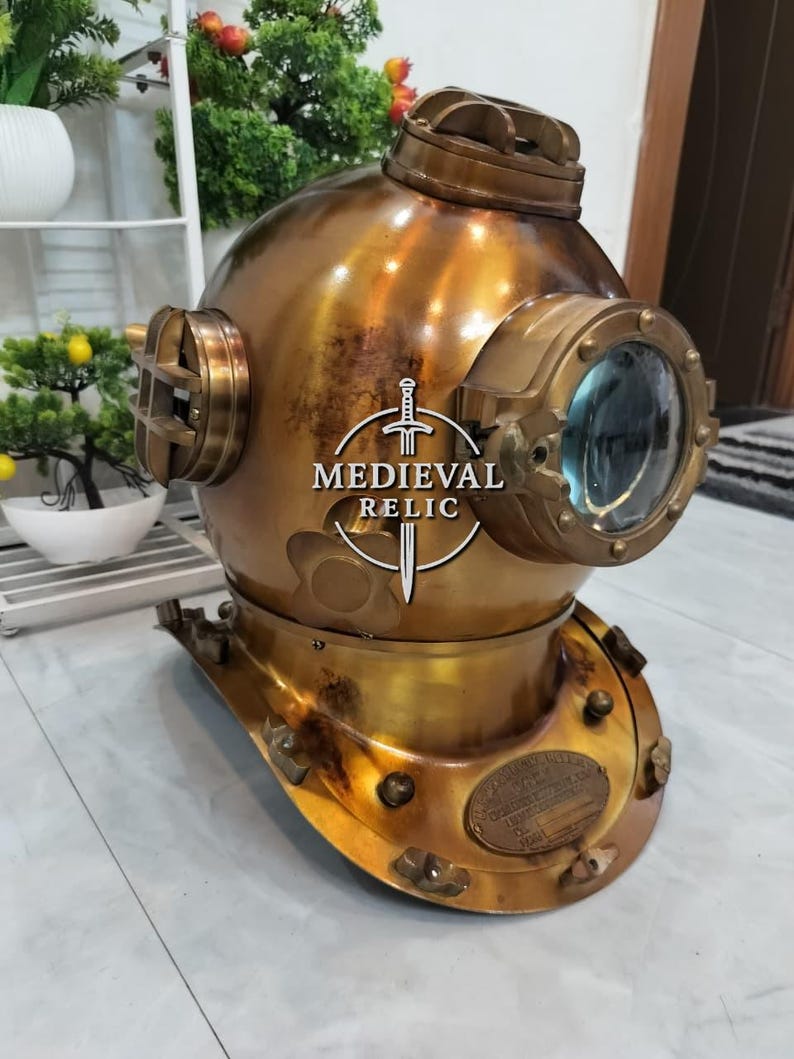 May include: A vintage brass diving helmet with a large front viewing port and side portholes. The helmet has a weathered, antique finish, and a plate with text is visible on the front. The helmet is a decorative item.