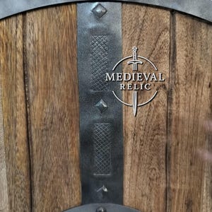 Puede incluir: Primer plano de un escudo de madera con detalles met&aacute;licos. El escudo tiene una parte superior curvada y est&aacute; reforzado con bandas y remaches met&aacute;licos. El centro presenta el texto "MEDIEVAL RELIC" con un gr&aacute;fico de espada.