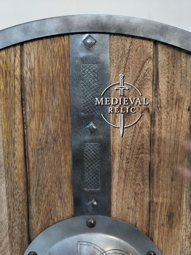 Puede incluir: Primer plano de un escudo de madera con detalles met&aacute;licos. El escudo tiene una parte superior curvada y est&aacute; reforzado con bandas y remaches met&aacute;licos. El centro presenta el texto "MEDIEVAL RELIC" con un gr&aacute;fico de espada.