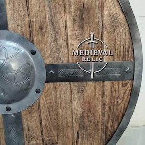 Puede incluir: Un escudo de madera con borde y detalles met&aacute;licos. El escudo tiene un dise&ntilde;o circular con un jefe central de metal y bandas horizontales de metal. El texto "MEDIEVAL RELIC" se muestra en el escudo.