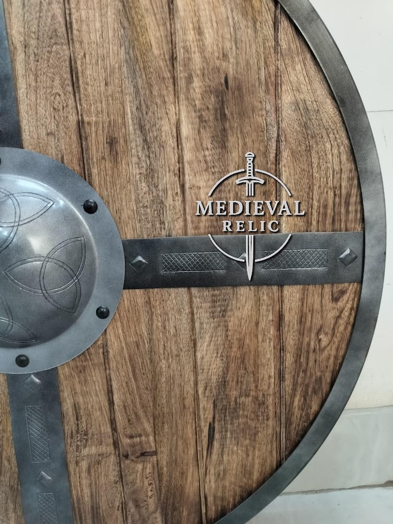 Puede incluir: Un escudo de madera con borde y detalles met&aacute;licos. El escudo tiene un dise&ntilde;o circular con un jefe central de metal y bandas horizontales de metal. El texto "MEDIEVAL RELIC" se muestra en el escudo.