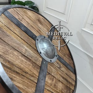 Puede incluir: Un escudo de madera redondo con borde y barras transversales de metal. El escudo tiene un jefe central de metal con un dise&ntilde;o de nudo celta en relieve. La madera tiene un veteado natural y el metal un aspecto envejecido.