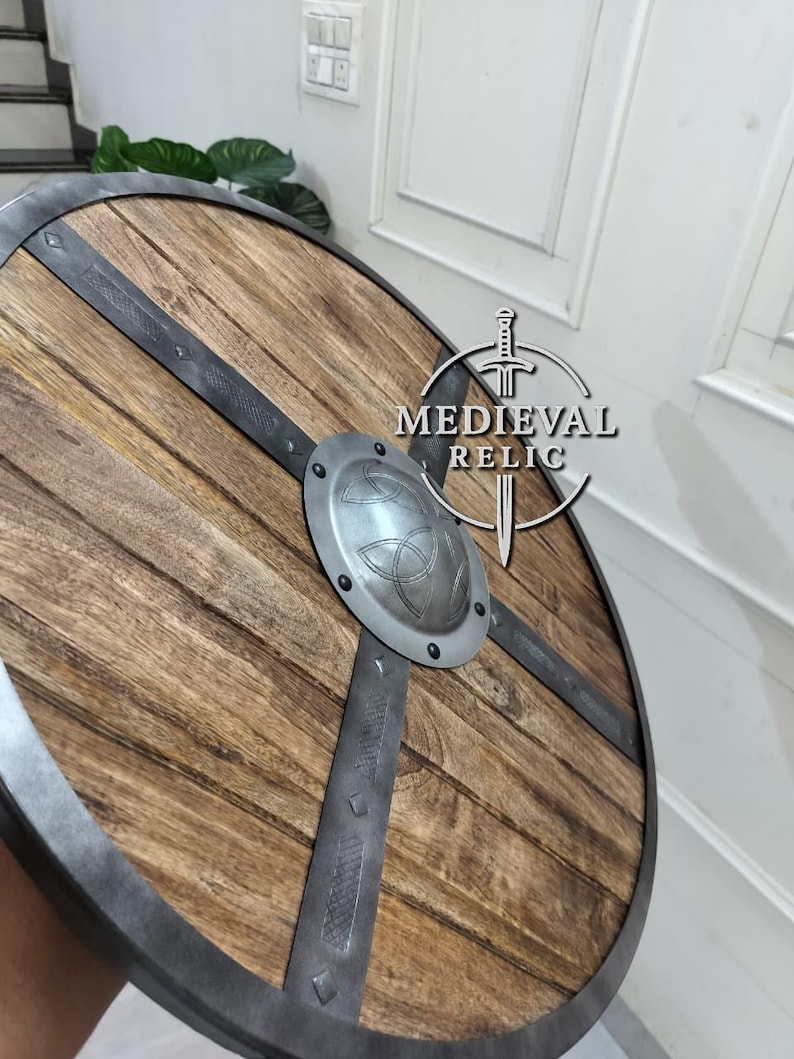 Puede incluir: Un escudo de madera redondo con borde y barras transversales de metal. El escudo tiene un jefe central de metal con un dise&ntilde;o de nudo celta en relieve. La madera tiene un veteado natural y el metal un aspecto envejecido.