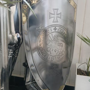 May include: A polished steel shield with a cross emblem and a circular seal featuring a knight on horseback. The shield has brass accents and is part of a suit of armor. The text "Medieval Relic" is visible.