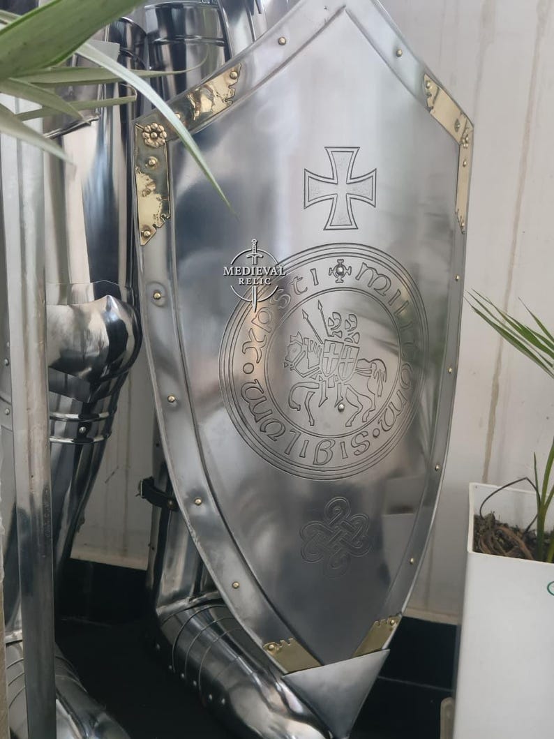 May include: A polished steel shield with a cross emblem and a circular seal featuring a knight on horseback. The shield has brass accents and is part of a suit of armor. The text "Medieval Relic" is visible.