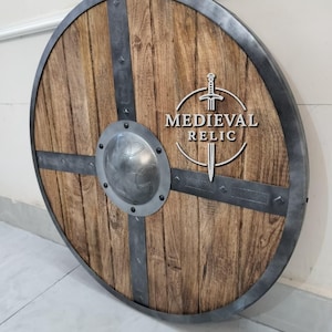 Puede incluir: Un escudo redondo de madera con borde y barras transversales de metal. El escudo tiene un aspecto de veta de madera envejecida con un centro met&aacute;lico. El texto "MEDIEVAL RELIC" est&aacute; visible.