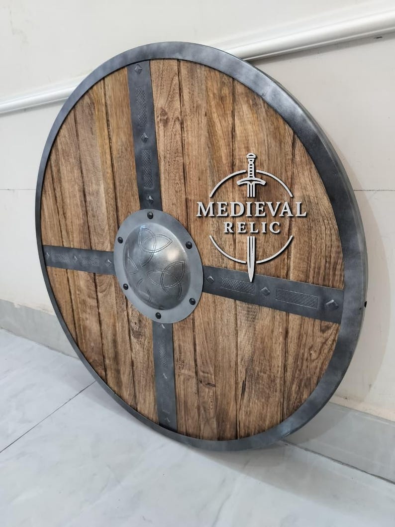 Puede incluir: Un escudo redondo de madera con borde y barras transversales de metal. El escudo tiene un aspecto de veta de madera envejecida con un centro met&aacute;lico. El texto "MEDIEVAL RELIC" est&aacute; visible.