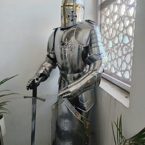 May include: A full suit of polished steel medieval armor, including a helmet, breastplate, and arm and leg protection. The armor is displayed with a sword and shield, and the shield features a cross design. The text "MEDIEVAL RELIC" is visible on the breastplate.