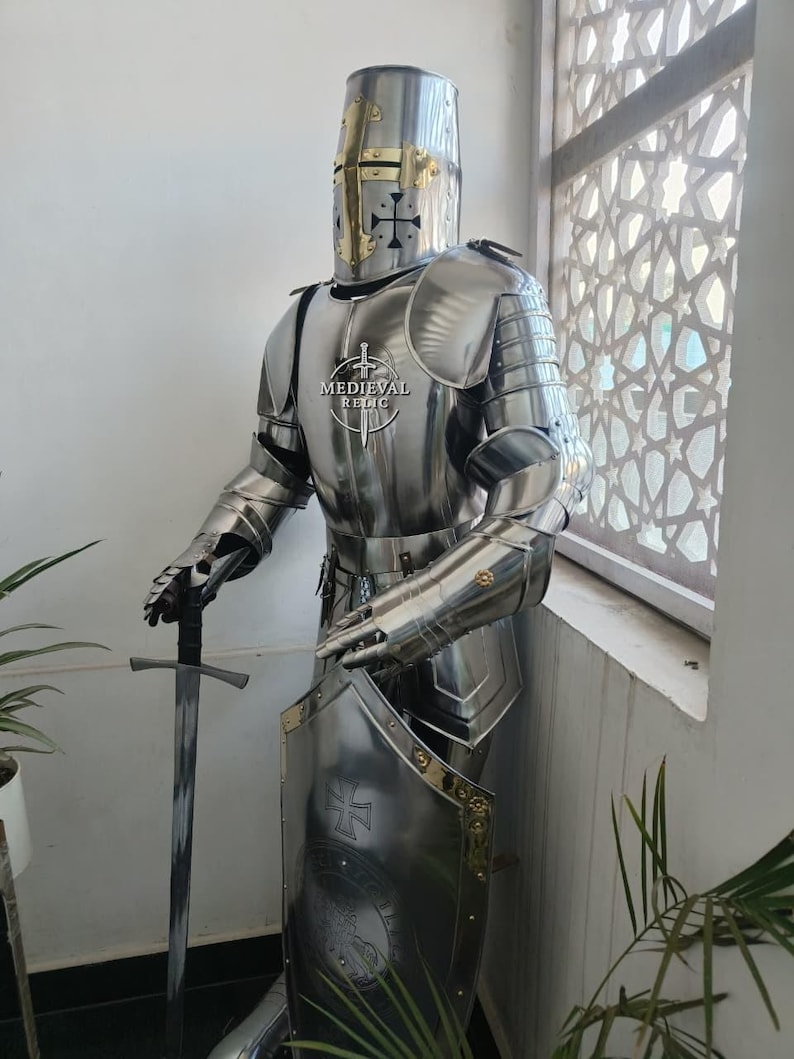May include: A full suit of polished steel medieval armor, including a helmet, breastplate, and arm and leg protection. The armor is displayed with a sword and shield, and the shield features a cross design. The text "MEDIEVAL RELIC" is visible on the breastplate.