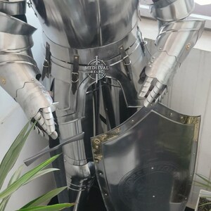May include: A polished, silver-colored suit of armor, including a breastplate, arm and hand protection, and leg coverings. A matching shield with gold trim is held in front. The armor has visible rivets and leather straps. The Medieval Relic logo is visible.