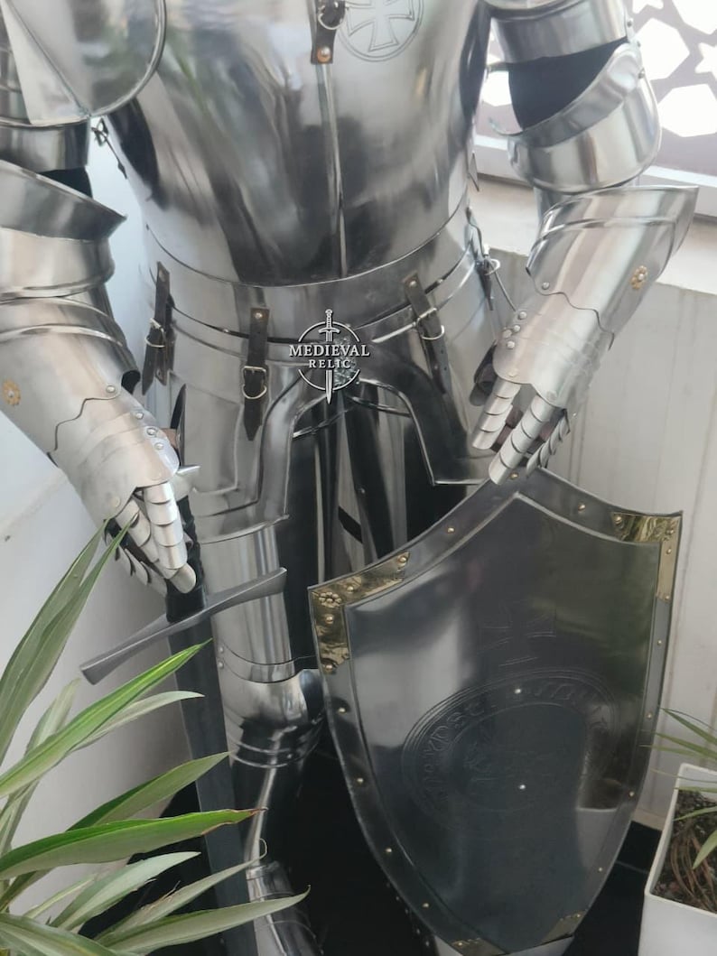May include: A polished, silver-colored suit of armor, including a breastplate, arm and hand protection, and leg coverings. A matching shield with gold trim is held in front. The armor has visible rivets and leather straps. The Medieval Relic logo is visible.