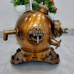 May include: A vintage brass diving helmet with the text "MEDIEVAL RELIC" and a sword emblem. The helmet has a round shape with multiple fittings, a viewing window, and a base with decorative elements. The brass has a warm, aged patina.