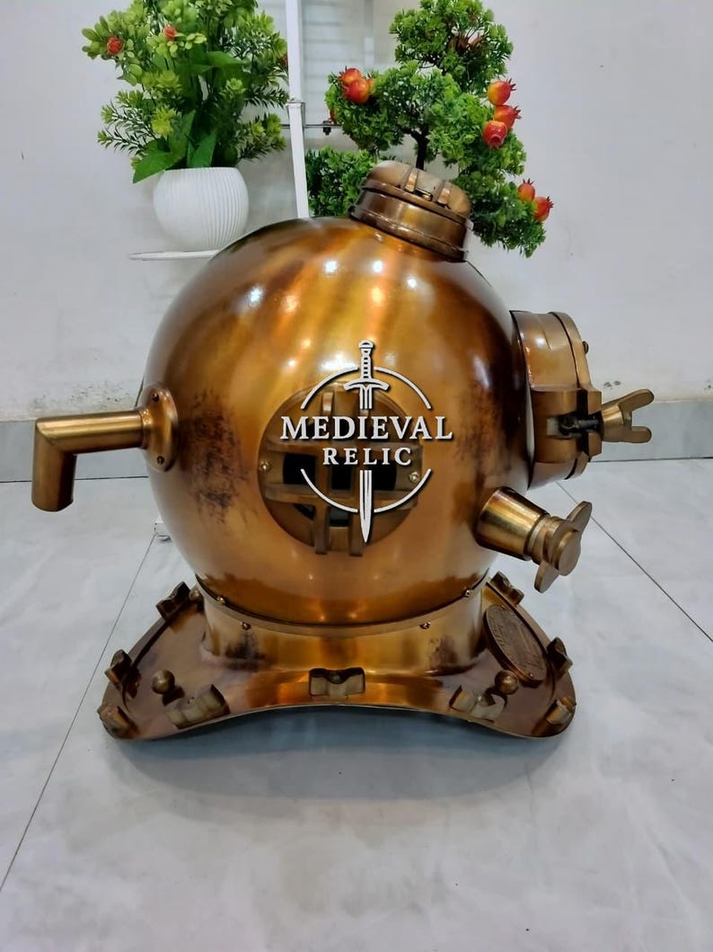 May include: A vintage brass diving helmet with the text "MEDIEVAL RELIC" and a sword emblem. The helmet has a round shape with multiple fittings, a viewing window, and a base with decorative elements. The brass has a warm, aged patina.